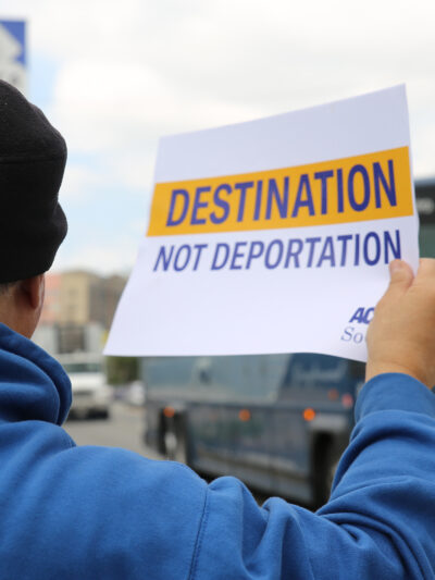 Man on sidewalk, holding a sign as a  Greyhound bus passes by: "Destination not deportation"