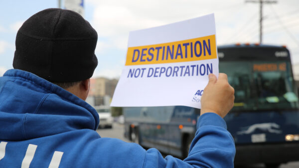 Man on sidewalk, holding a sign as a  Greyhound bus passes by: "Destination not deportation"