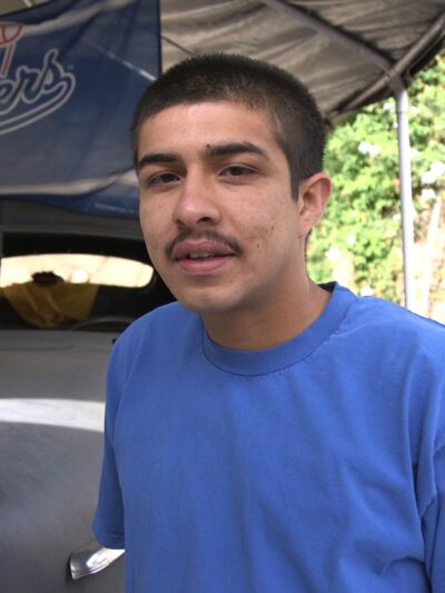 Peter Arrellano, plaintiff in YJC v. Los Angeles, standing in front of a Volkswagen bug in a carport with a Dodgers flag hanging from the top of the carport