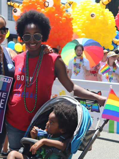 Two women with a child in a stroller in the foreground at a Pride Parade. One woman is holding a sign that reads We the People.
