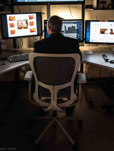 A man sitting in front of 3 computer screens, with more surrounding him, using the equipment for surveillance.