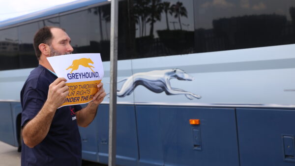 A man on the sidewalk holding a sign that reads "Greyhound, stop throwing our rights under the bus" as a Greyhound bus passes by.