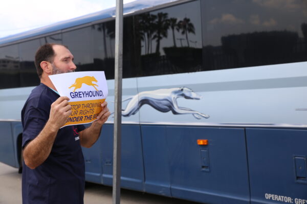 A man on the sidewalk holding a sign that reads "Greyhound, stop throwing our rights under the bus" as a Greyhound bus passes by.