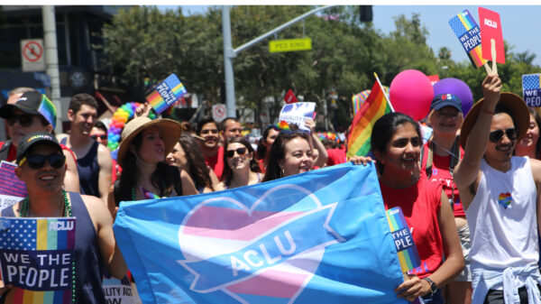 Annual Report 2018 ACLU of Southern California. Image of a group of people marching in the Los Angeles Pride Parade, the people in the front holding a blue flag with a pink and blue ACLU heart on it.