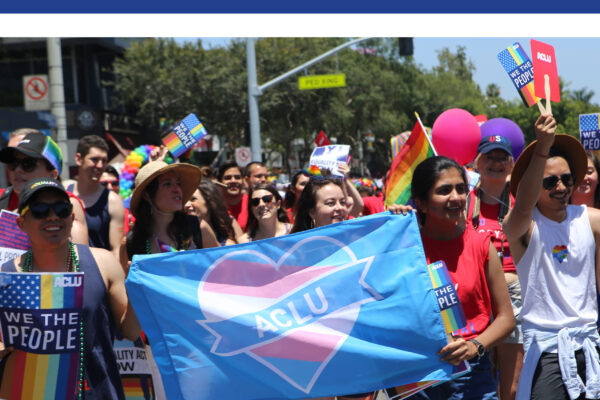 Annual Report 2018 ACLU of Southern California. Image of a group of people marching in the Los Angeles Pride Parade, the people in the front holding a blue flag with a pink and blue ACLU heart on it.