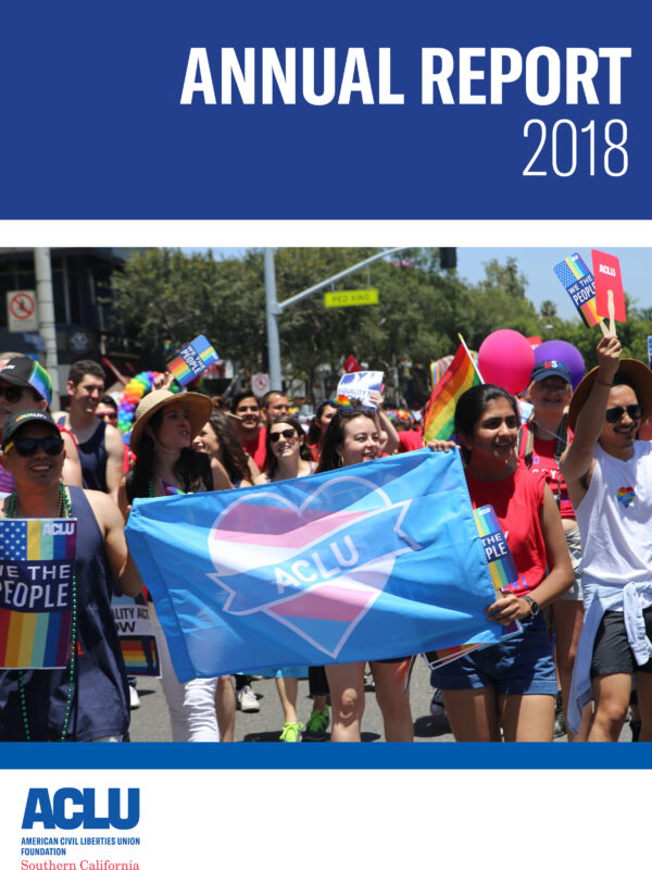 Annual Report 2018 ACLU of Southern California. Image of a group of people marching in the Los Angeles Pride Parade, the people in the front holding a blue flag with a pink and blue ACLU heart on it.