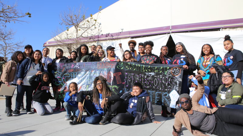 A group of high school students gathered around a hand-written banner that reads: students not suspects