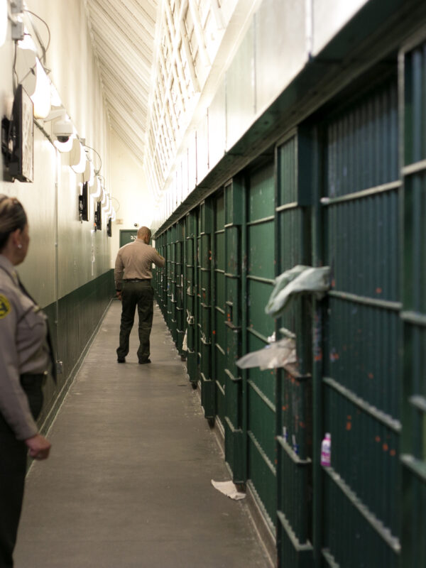 A female sheriff's deputy standing in front of a jail cell, she's looking to her left where a male sheriff's deputy is walking down the hall with his back to her.