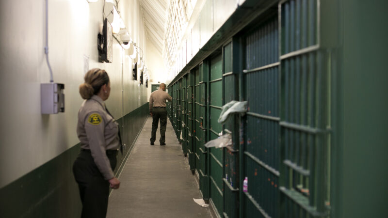 A female sheriff's deputy standing in front of a jail cell, she's looking to her left where a male sheriff's deputy is walking down the hall with his back to her.