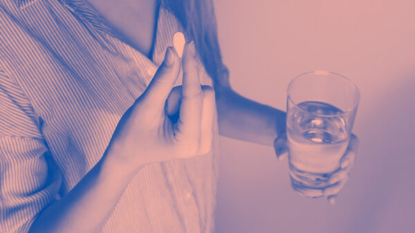 A woman holding a white pill in her right hand and a glass of water in her left hand.
