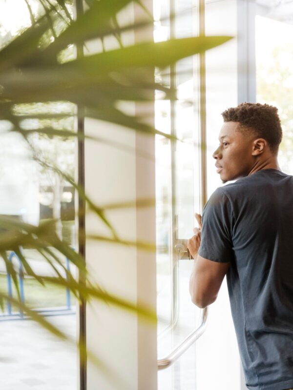 In the background, a young man with a backpack opening and walking through a glass door. In the foreground, the leaves of a palm tree.