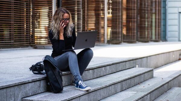 A young woman with shoulder length blonde hair, sitting on the steps of a large building, with a cell phone to her ear and a laptop propped on her knees, a backpack next to her on the ground.