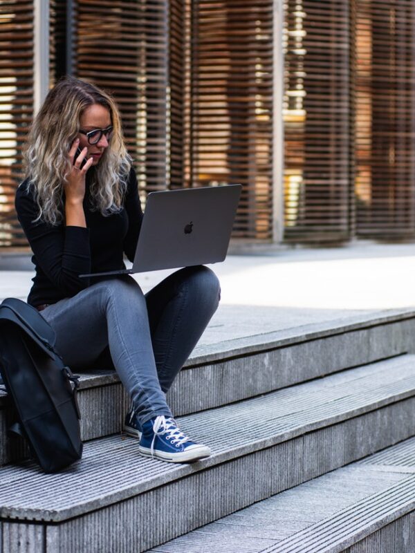 A young woman with shoulder length blonde hair, sitting on the steps of a large building, with a cell phone to her ear and a laptop propped on her knees, a backpack next to her on the ground.