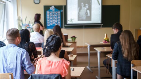 A school classroom with a teacher at standing at the blackboard with a projector screen and students sitting at their desks.