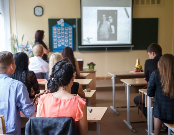 A school classroom with a teacher at standing at the blackboard with a projector screen and students sitting at their desks.