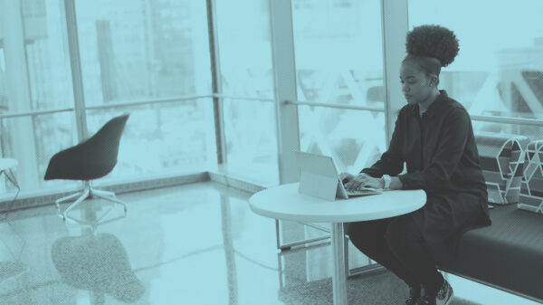 A young Black woman with an afro puff sitting on a sofa, using her laptop at a small table. Image courtesy of #WOCinTech Chat.