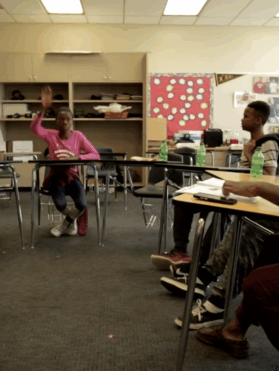 Six youth and an adult sitting in a classroom with their desks forming a semi-circle. Three of the youth have their hands raised.