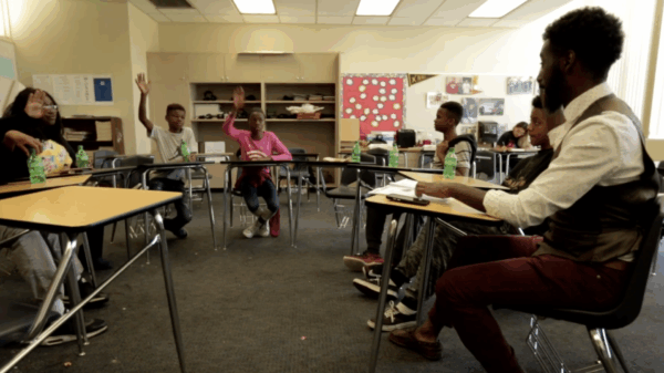 Six youth and an adult sitting in a classroom with their desks forming a semi-circle. Three of the youth have their hands raised.