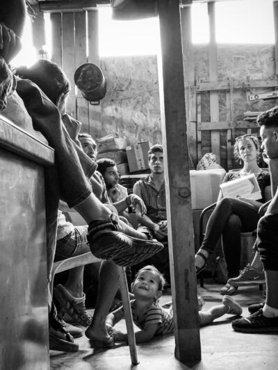 Ana Adlerstein sitting with a small group of Central American migrants in a room, a toddler is crawling on the floor looking up at a seated adult