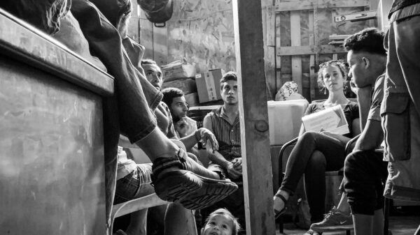 Ana Adlerstein sitting with a small group of Central American migrants in a room, a toddler is crawling on the floor looking up at a seated adult