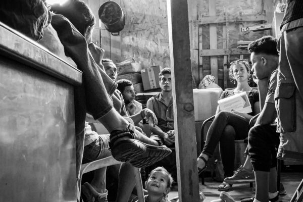Ana Adlerstein sitting with a small group of Central American migrants in a room, a toddler is crawling on the floor looking up at a seated adult