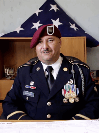 Hector Barajas wearing in his U.S. Marine uniform seated at his desk, an American flag folded into a triangle on a shelf behind him