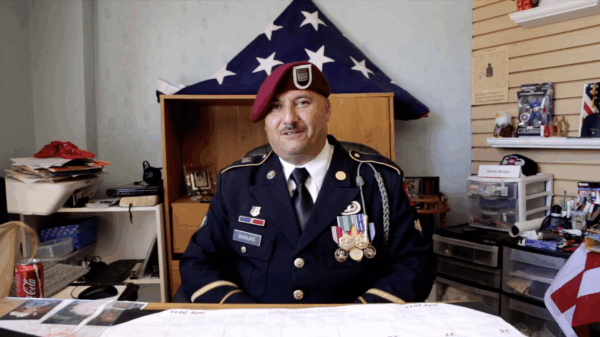 Hector Barajas wearing in his U.S. Marine uniform seated at his desk, an American flag folded into a triangle on a shelf behind him