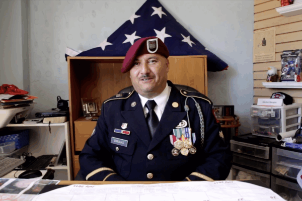 Hector Barajas wearing in his U.S. Marine uniform seated at his desk, an American flag folded into a triangle on a shelf behind him