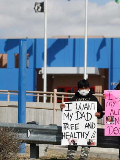 Two children wearing face masks standing in front of an immigration detention center holding signs that read: "I want my dad free and healthy" and "I want my dad alive not dead."