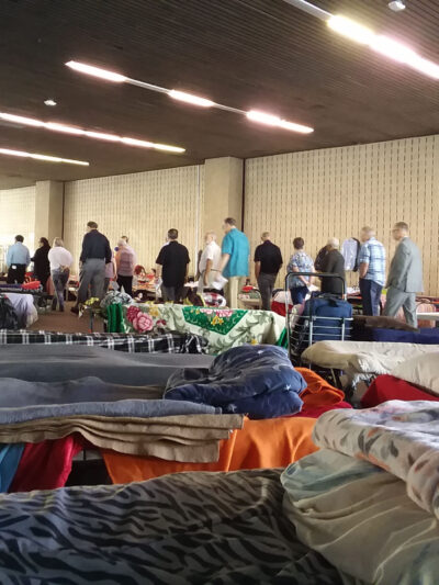 A scene from a homeless shelter in Orange County. In the foreground, cots with blankets and pillows a couple feet apart. In the background, a group of people standing in a line.