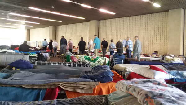 A scene from a homeless shelter in Orange County. In the foreground, cots with blankets and pillows a couple feet apart. In the background, a group of people standing in a line.
