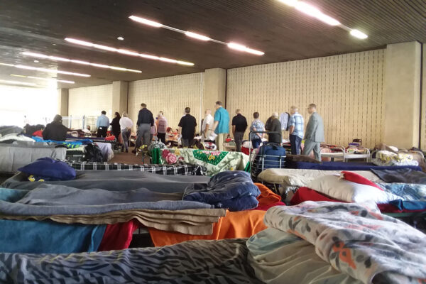 A scene from a homeless shelter in Orange County. In the foreground, cots with blankets and pillows a couple feet apart. In the background, a group of people standing in a line.