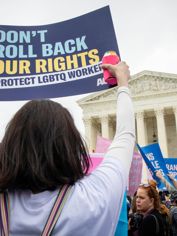 A person at a protest rally in front of the U.S. Supreme court holding a sign that reads: Don't roll back our rights. Protect LGBTQ workers.