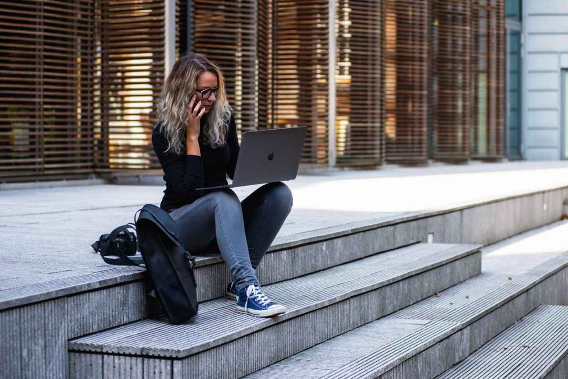 A young woman with shoulder length blonde hair, sitting on the steps of a large building, with a cell phone to her ear and a laptop propped on her knees, a backpack next to her on the ground.