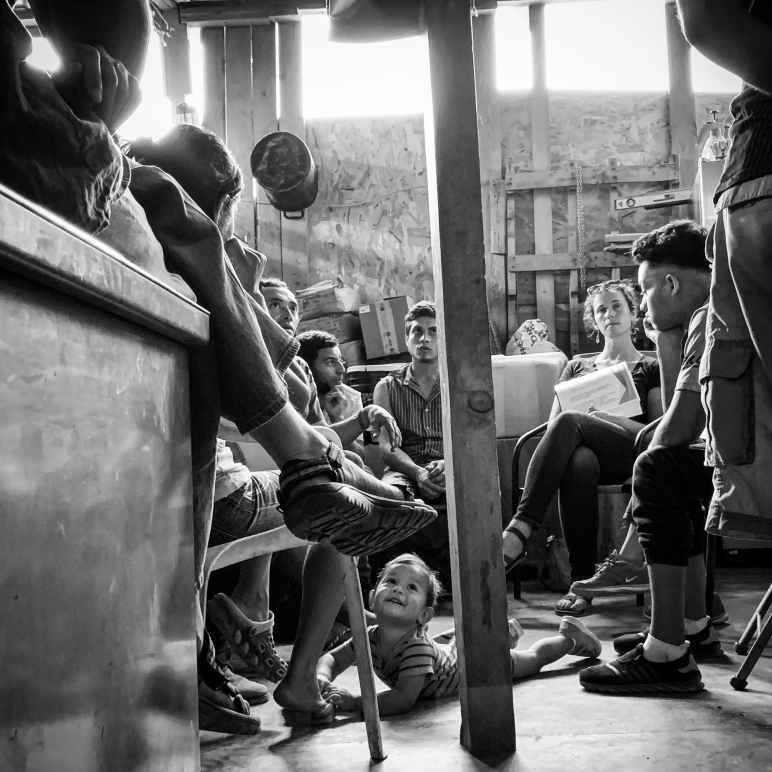 Ana Adlerstein sitting with a small group of Central American migrants in a room, a toddler is crawling on the floor looking up at a seated adult