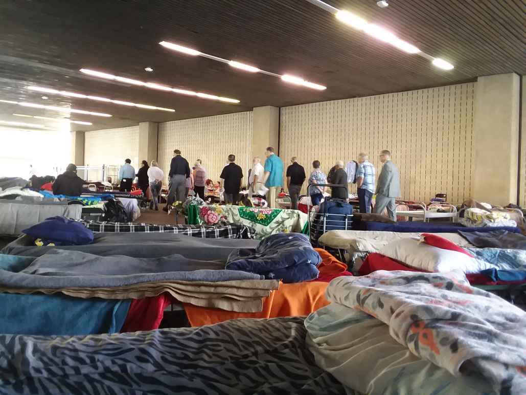 A scene from a homeless shelter in Orange County. In the foreground, cots with blankets and pillows a couple feet apart. In the background, a group of people standing in a line.