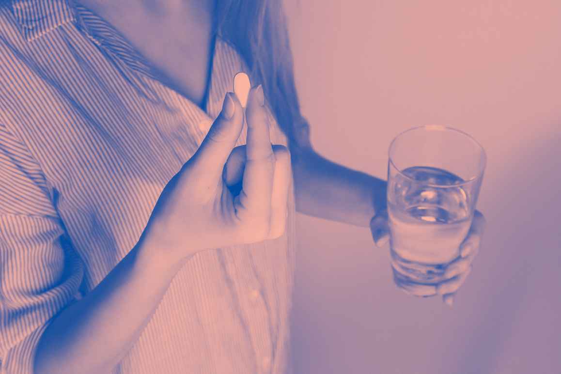 A woman holding a white pill in her right hand and a glass of water in her left hand.