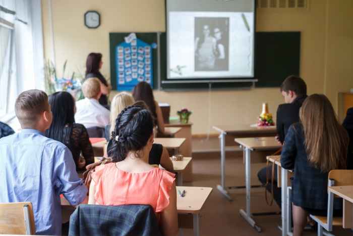 A school classroom with a teacher at standing at the blackboard with a projector screen and students sitting at their desks.