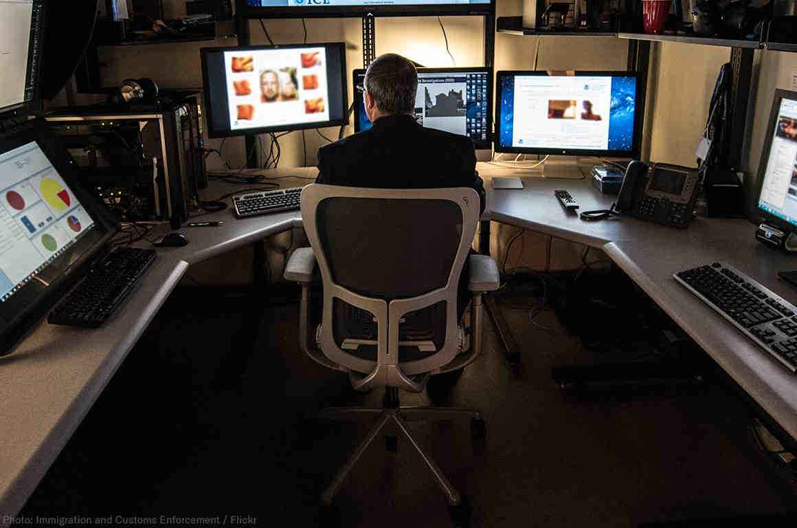 A man sitting in front of 3 computer screens, with more surrounding him, using the equipment for surveillance.