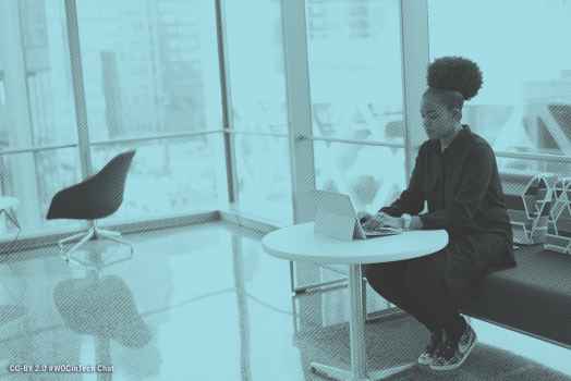 A young Black woman with an afro puff sitting on a sofa, using her laptop at a small table. Image courtesy of #WOCinTech Chat.