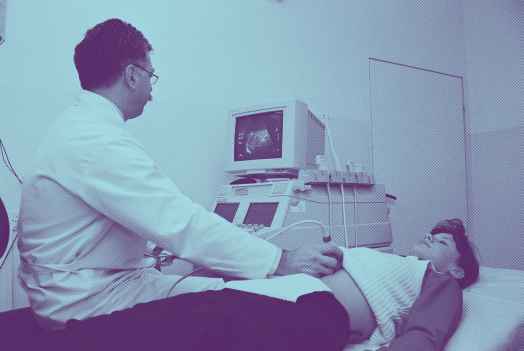 A woman lying on an exam table, a male doctor performing an ultrasound by placing a wand over her abdominal area