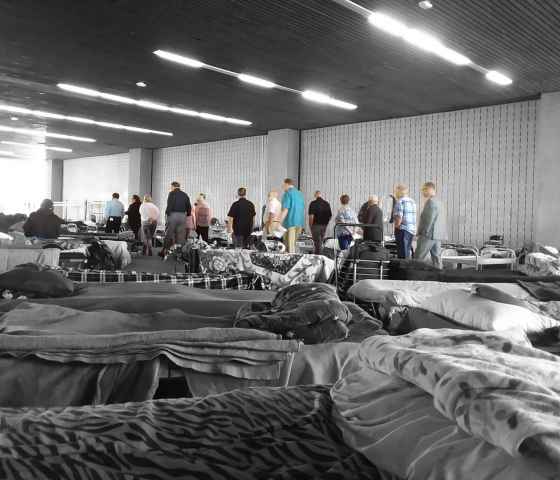 A scene from a homeless shelter in Orange County. In the foreground, cots with blankets and pillows a couple feet apart. In the background, a group of people standing in a line.