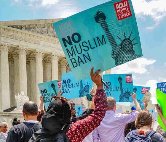 A woman wearing a hijab holding a sign that reads: No Muslim Ban